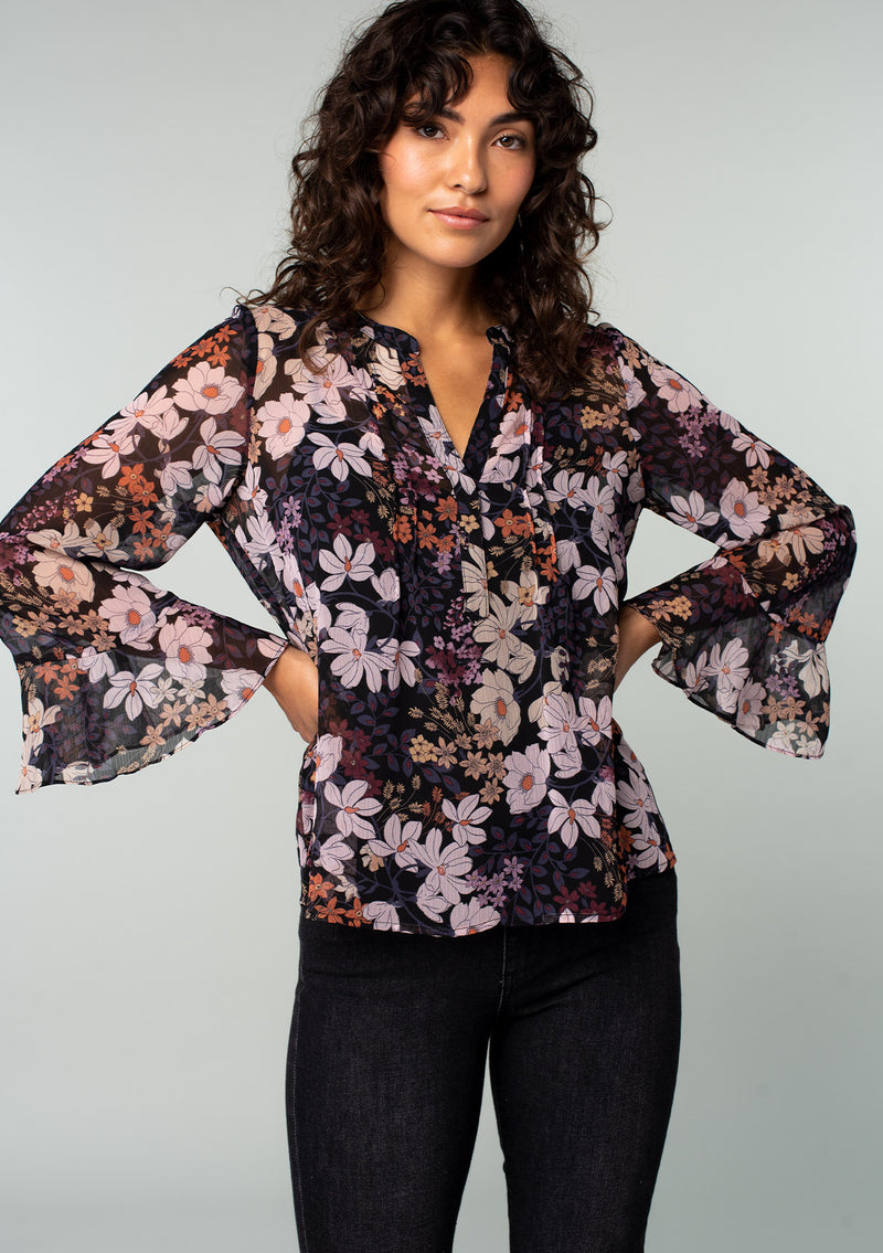 [Color: Black/Dusty Rose] A front facing image of a brunette model wearing a sheer chiffon bohemian blouse in a black and rose pink floral print. With long sleeves, ruffled wrist cuffs, and a split v neckline. 