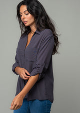 [Color: Charcoal] A half body side image of a brunette model wearing a relaxed fit charcoal grey button front shirt in a lightweight crepe. With long rolled sleeves, a button tab sleeve closure, a front placket, and a collared neckline. 
