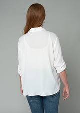 [Color: Gardenia] A back facing image of a red headed model wearing a relaxed fit ivory white button front shirt in a lightweight crepe. With long rolled sleeves, a button tab sleeve closure, a front placket, and a collared neckline.