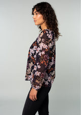 [Color: Black/Dusty Rose] A side facing image of a brunette model wearing a chiffon bohemian blouse in a black and purple floral print. With long sleeves and a smocked elastic round neckline.