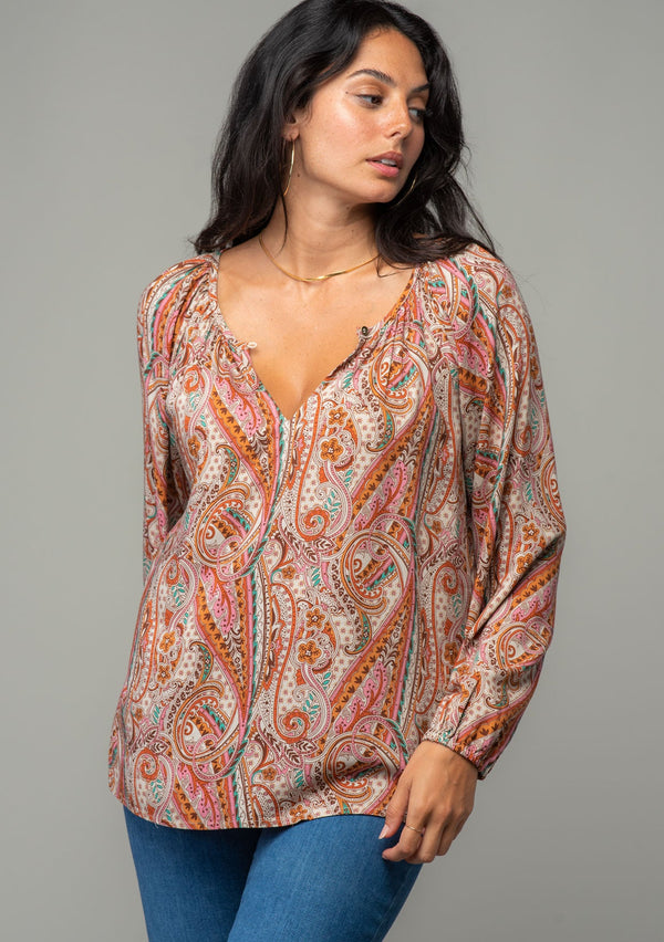 [Color: Natural/Rust] A front facing image of a brunette model wearing a bohemian peasant top in a natural and rust red paisley print. With long sleeves, a relaxed flowy fit, and a button loop closure at the neckline. 