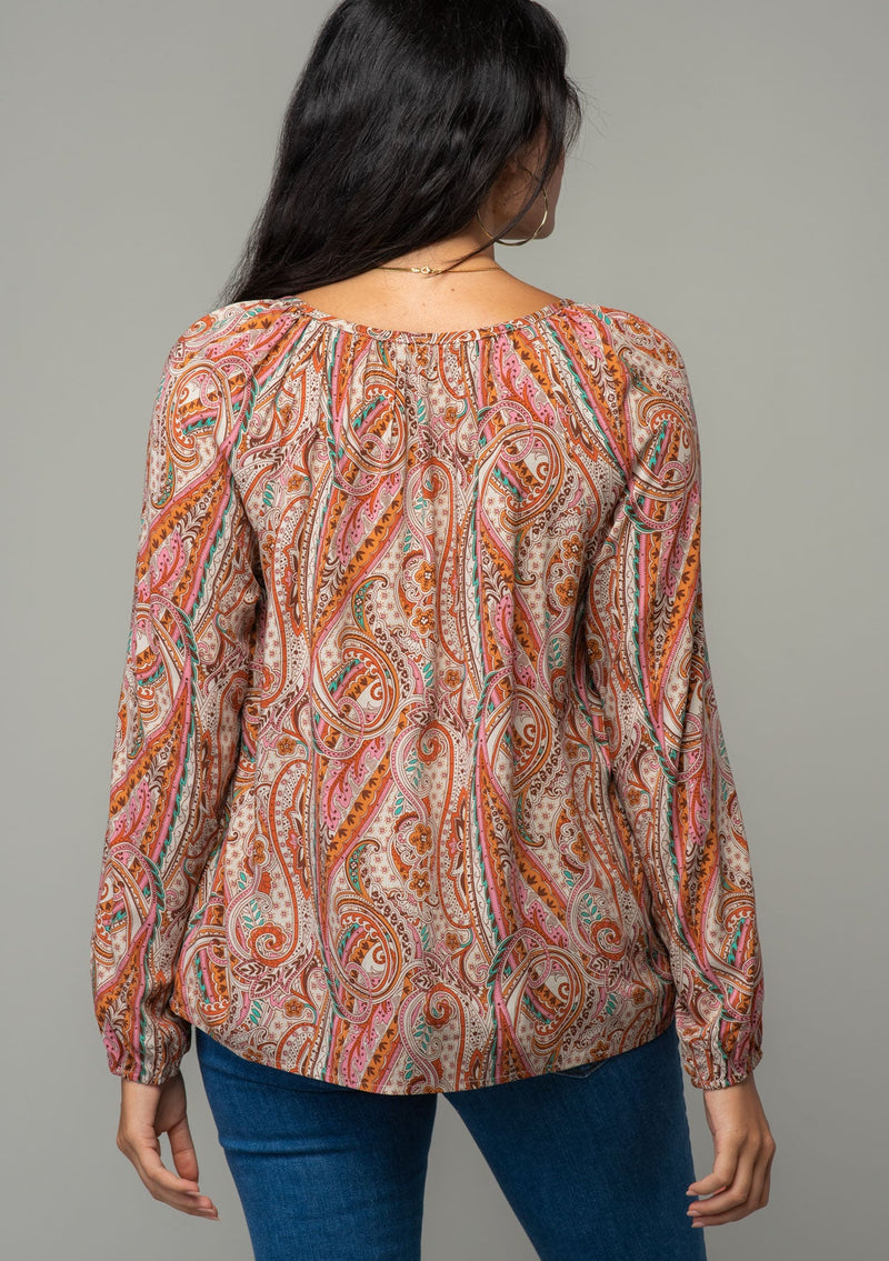[Color: Natural/Rust] A back facing image of a brunette model wearing a bohemian peasant top in a natural and rust red paisley print. With long sleeves, a relaxed flowy fit, and a button loop closure at the neckline. 
