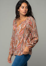 [Color: Natural/Rust] A side facing image of a brunette model wearing a bohemian peasant top in a natural and rust red paisley print. With long sleeves, a relaxed flowy fit, and a button loop closure at the neckline. 