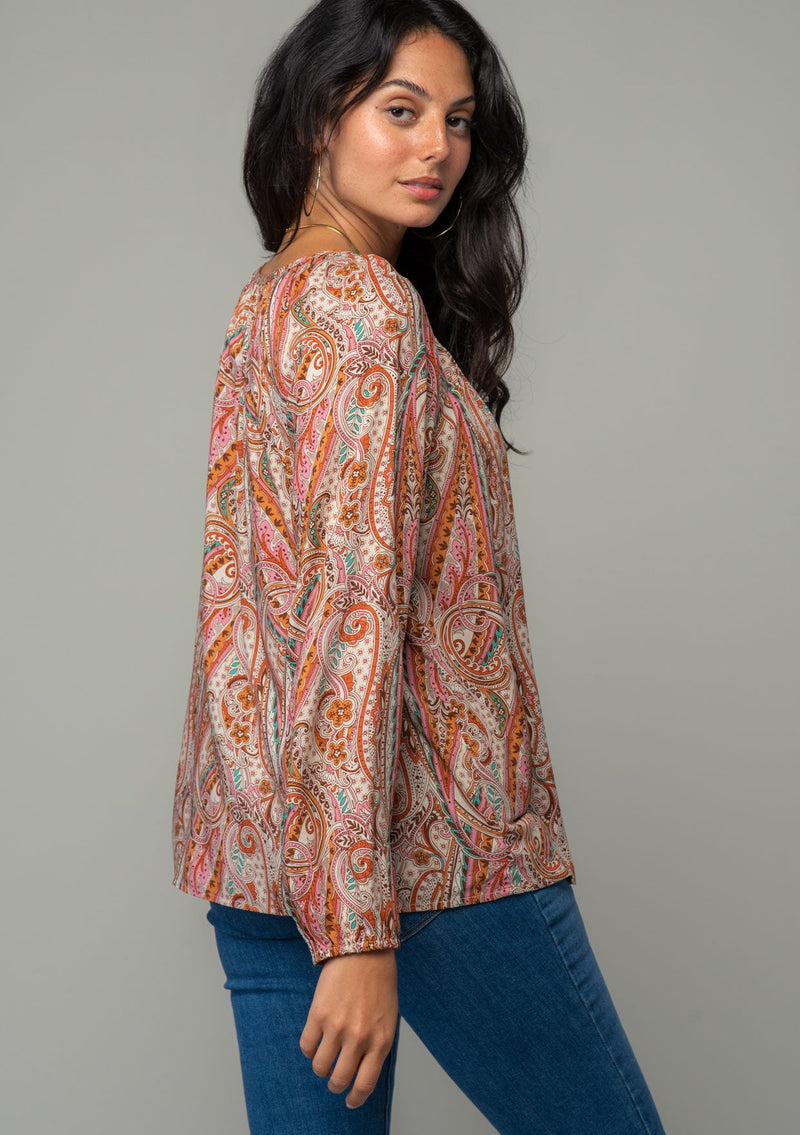 [Color: Natural/Rust] A back facing image of a brunette model wearing a bohemian peasant top in a natural and rust red paisley print. With long sleeves, a relaxed flowy fit, and a button loop closure at the neckline. The model is looking over her shoulder. 
