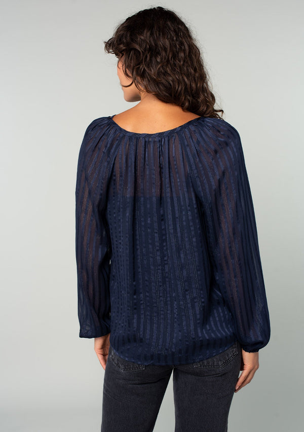 [Color: Navy] A back facing image of a brunette model wearing a sheer bohemian navy blue holiday blouse in a sparkly lurex stripe. With voluminous long sleeves and a v neckline. 
