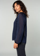 [Color: Navy] A side facing image of a brunette model wearing a sheer bohemian navy blue holiday blouse in a sparkly lurex stripe. With voluminous long sleeves and a v neckline. 