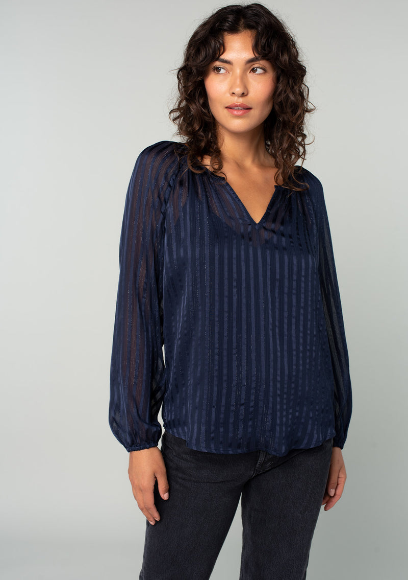 [Color: Navy] A front facing image of a brunette model wearing a sheer bohemian navy blue holiday blouse in a sparkly lurex stripe. With voluminous long sleeves and a v neckline. 