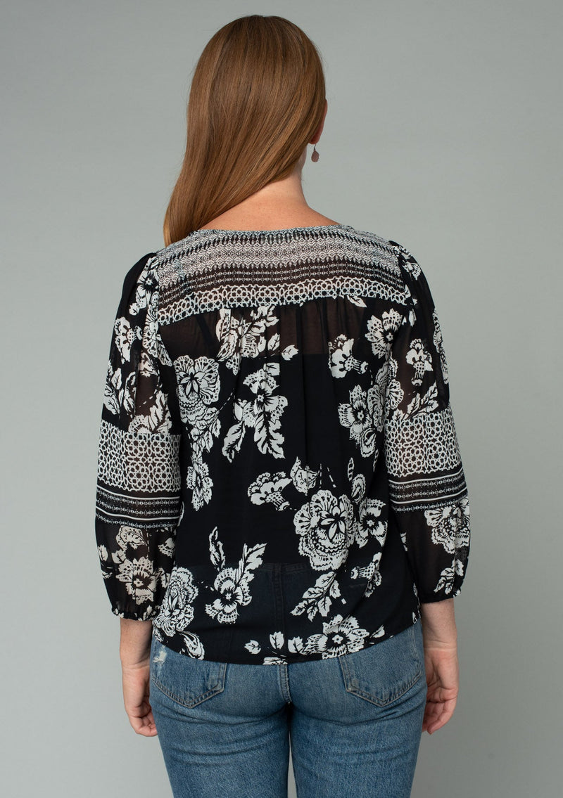 [Color: Black/Cream] A back facing image of a red headed model wearing a bohemian chiffon blouse in a black and cream mixed floral print. With a button front and three quarter length sleeves. 