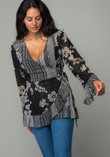 [Color: Black/Cream] A front facing image of a brunette model wearing a bohemian hippie wrap top in a mixed black and cream floral print. With long bell sleeves and a ruffled wrist cuff, a deep v neckline, and a side tie closure. 