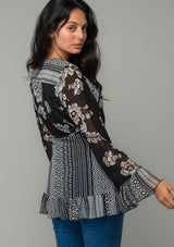 [Color: Black/Cream] A back facing image of a brunette model wearing a bohemian hippie wrap top in a mixed black and cream floral print. With long bell sleeves and a ruffled wrist cuff, a deep v neckline, and a side tie closure. 