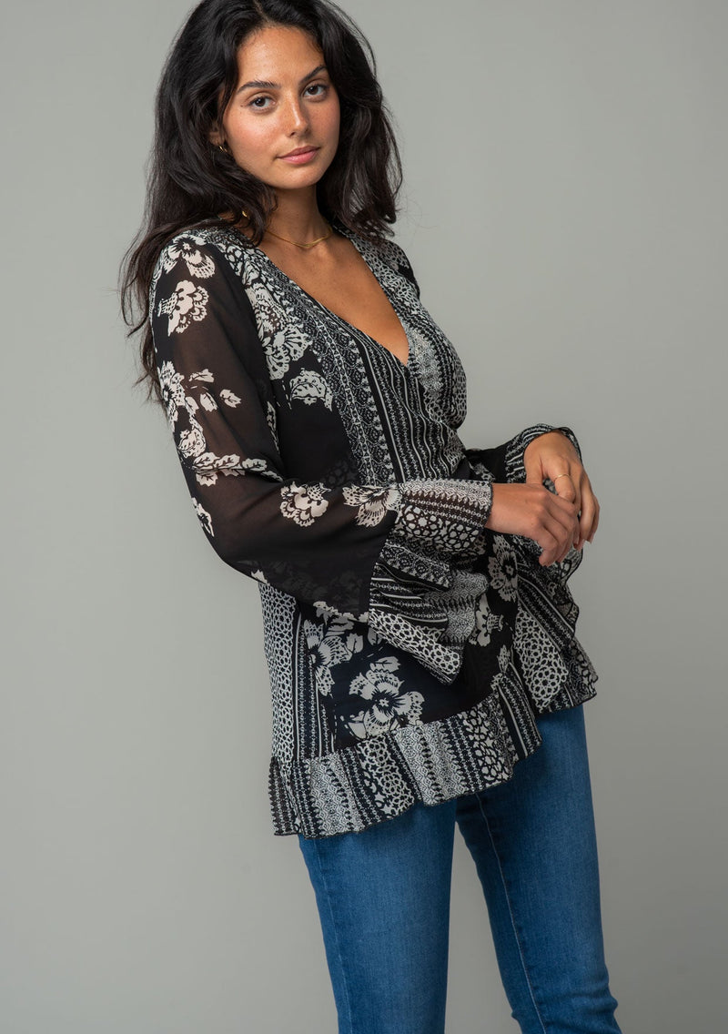 [Color: Black/Cream] A side facing image of a brunette model wearing a bohemian hippie wrap top in a mixed black and cream floral print. With long bell sleeves and a ruffled wrist cuff, a deep v neckline, and a side tie closure. 