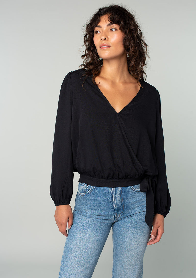 [Color: Black] A front facing image of a brunette model wearing a black bohemian holiday top with long sleeves, a faux wrap front, a surplice v neckline, and a side tie detail. 