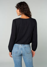 [Color: Black] A back facing image of a brunette model wearing a black bohemian holiday top with long sleeves, a faux wrap front, a surplice v neckline, and a side tie detail. 