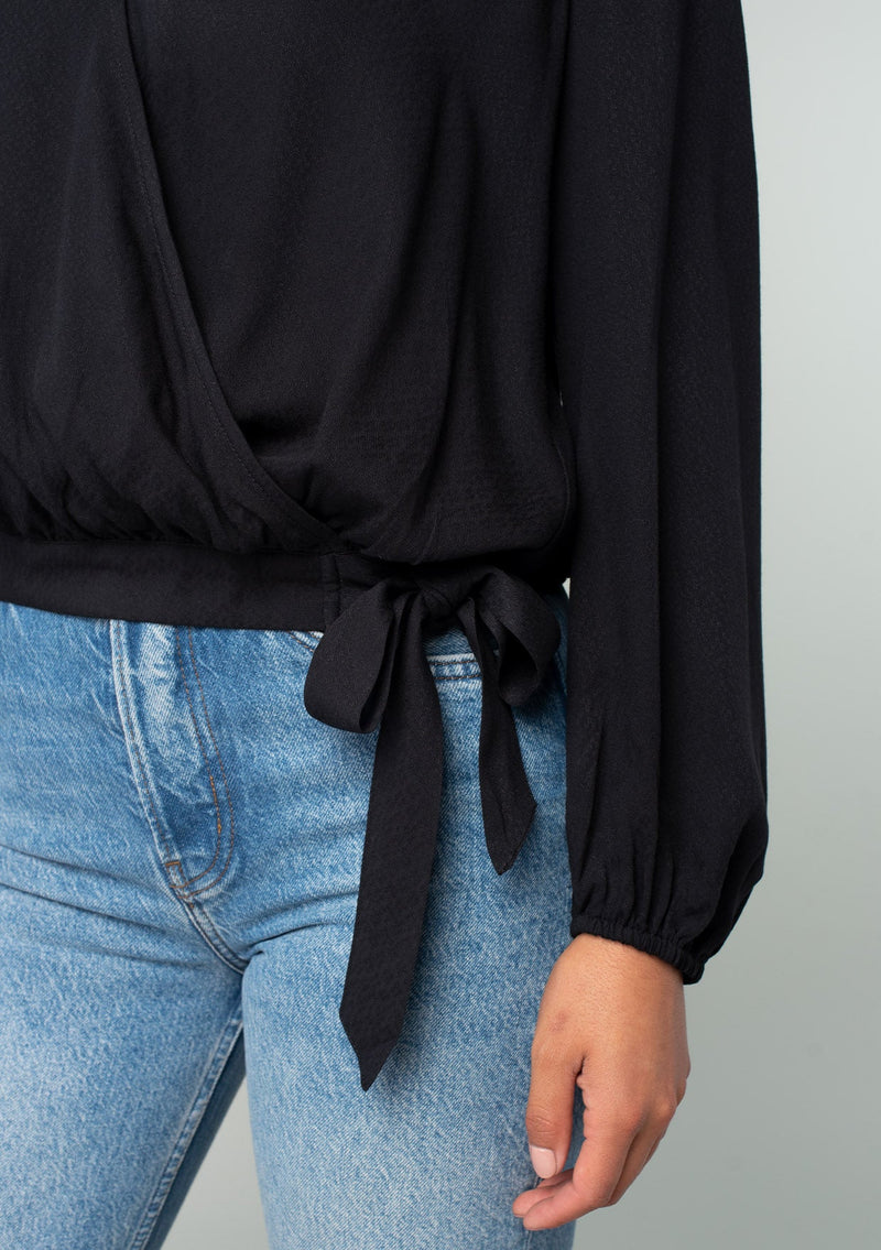 [Color: Black] A close up front facing image of a brunette model wearing a black bohemian holiday top with long sleeves, a faux wrap front, a surplice v neckline, and a side tie detail. 