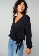 [Color: Black] A side facing image of a brunette model wearing a black bohemian holiday top with long sleeves, a faux wrap front, a surplice v neckline, and a side tie detail. 