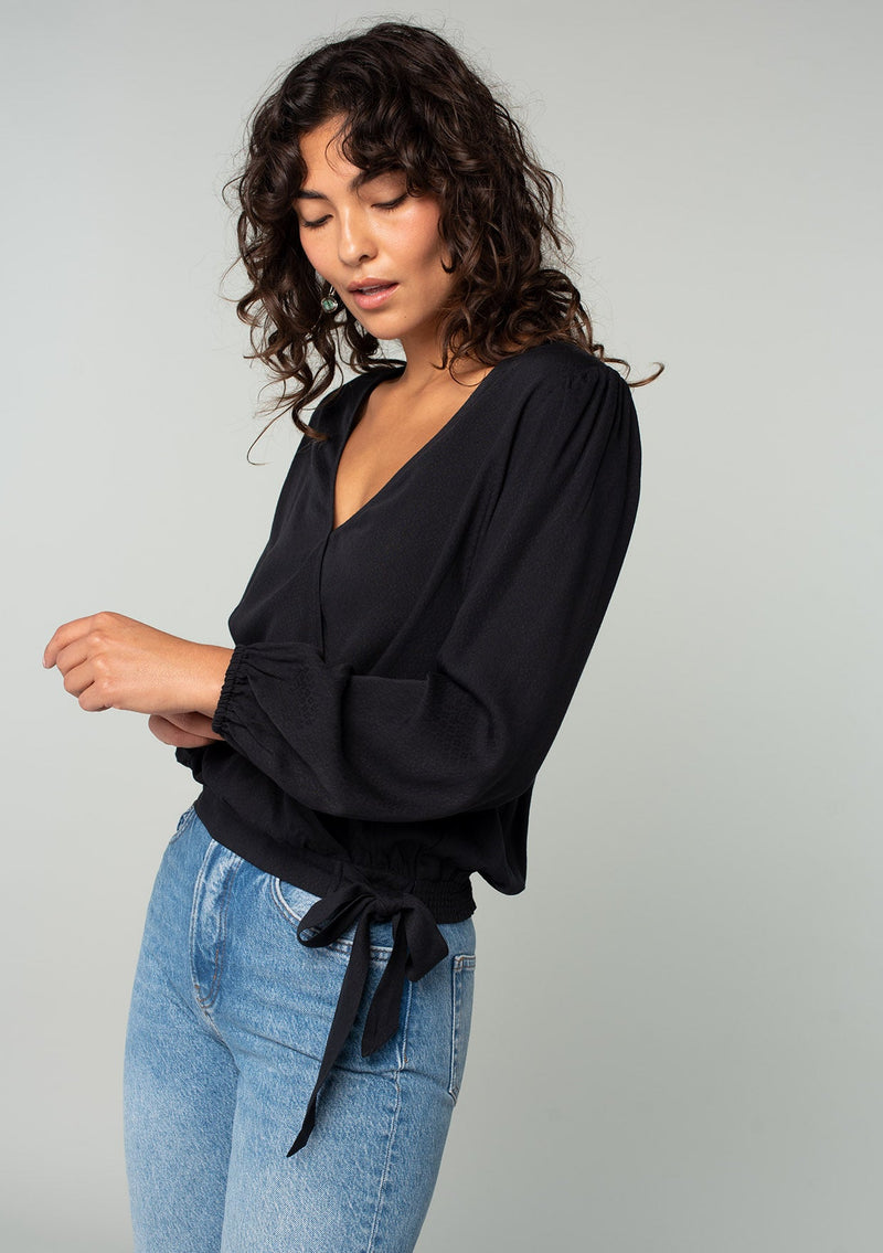 [Color: Black] A half body side facing image of a brunette model wearing a black bohemian holiday top with long sleeves, a faux wrap front, a surplice v neckline, and a side tie detail. 