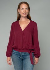 [Color: Wine] A half body front facing image of a red headed model wearing a wine red bohemian holiday top with long sleeves, a faux wrap front, a surplice v neckline, and a side tie detail. 