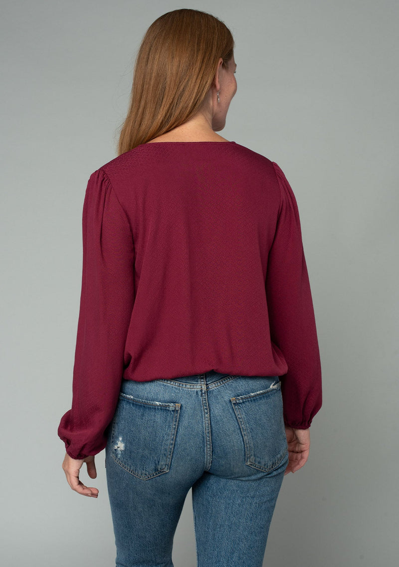 [Color: Wine] A back facing image of a red headed model wearing a wine red bohemian holiday top with long sleeves, a faux wrap front, a surplice v neckline, and a side tie detail. 
