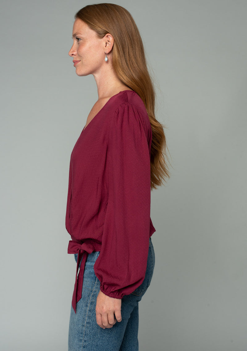 [Color: Wine] A side facing image of a red headed model wearing a wine red bohemian holiday top with long sleeves, a faux wrap front, a surplice v neckline, and a side tie detail. 