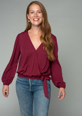 [Color: Wine] A front facing image of a red headed model wearing a wine red bohemian holiday top with long sleeves, a faux wrap front, a surplice v neckline, and a side tie detail. 