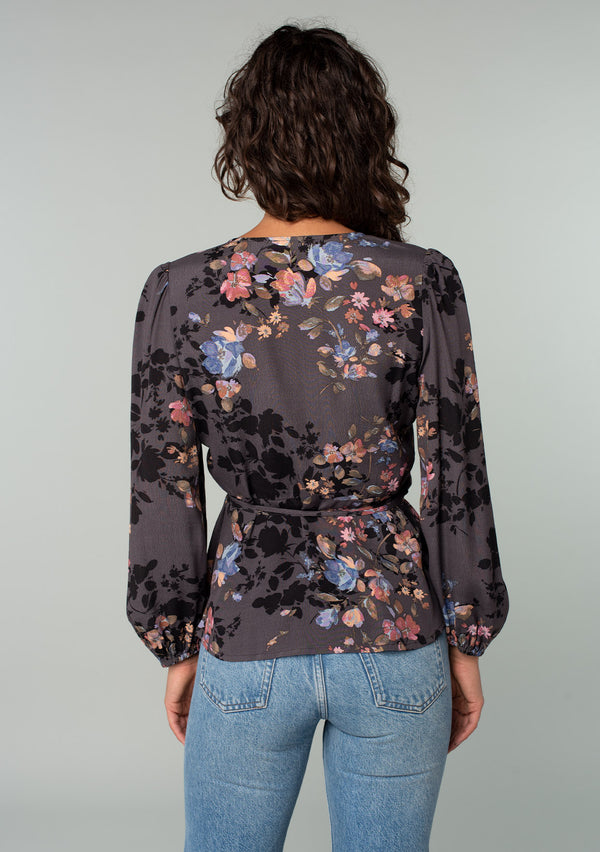 [Color: Grey/Dusty Blue] A back facing image of a model wearing a bohemian wrap top in a dark grey and dusty blue floral print. With long sleeves and a side tie closure. 
