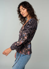 [Color: Grey/Dusty Blue] A side facing image of a model wearing a bohemian wrap top in a dark grey and dusty blue floral print. With long sleeves and a side tie closure. 