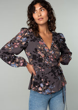 [Color: Grey/Dusty Blue] A front facing image of a model wearing a bohemian wrap top in a dark grey and dusty blue floral print. With long sleeves and a side tie closure. 