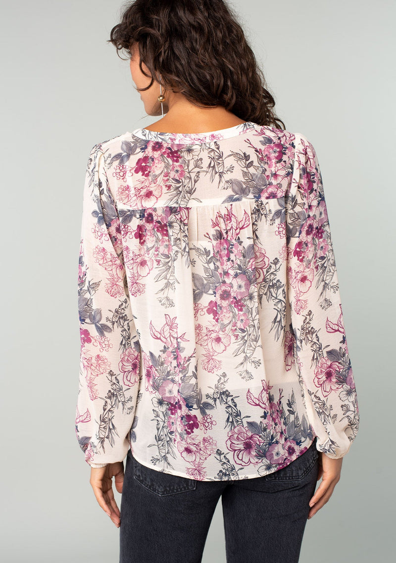 [Color: Natural/Wine] A back facing image of a brunette model wearing a sheer chiffon bohemian blouse in a natural and wine purple floral print. With a front placket, long voluminous sleeves, and a flowy relaxed fit. 