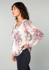 [Color: Natural/Wine] A side facing image of a brunette model wearing a sheer chiffon bohemian blouse in a natural and wine purple floral print. With a front placket, long voluminous sleeves, and a flowy relaxed fit. 