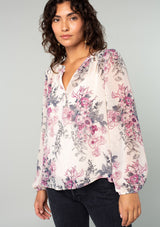[Color: Natural/Wine] A front facing image of a brunette model wearing a sheer chiffon bohemian blouse in a natural and wine purple floral print. With a front placket, long voluminous sleeves, and a flowy relaxed fit. 