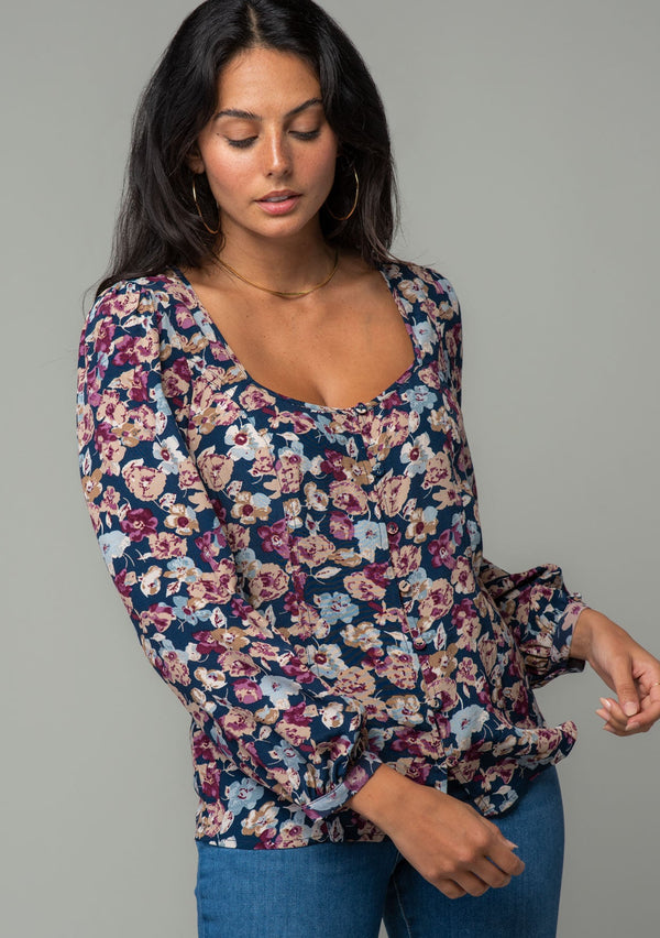 [Color: Teal/Plum] A front facing image of a brunette model wearing a bohemian blouse in a teal blue and plum purple floral print. With voluminous long sleeves, a self covered button up front, and a slight sweetheart neckline. 