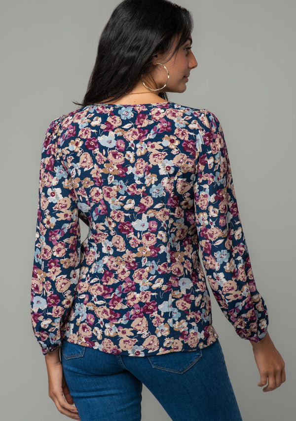 [Color: Teal/Plum] A back facing image of a brunette model wearing a bohemian blouse in a teal blue and plum purple floral print. With voluminous long sleeves, a self covered button up front, and a slight sweetheart neckline. 