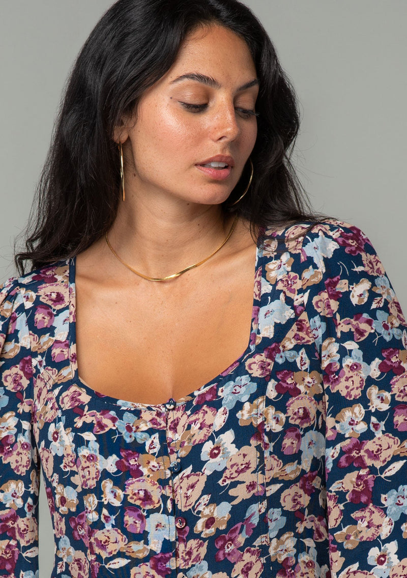 [Color: Teal/Plum] A close up front facing image of a brunette model wearing a bohemian blouse in a teal blue and plum purple floral print. With voluminous long sleeves, a self covered button up front, and a slight sweetheart neckline. 