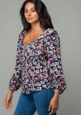 [Color: Teal/Plum] A side facing image of a brunette model wearing a bohemian blouse in a teal blue and plum purple floral print. With voluminous long sleeves, a self covered button up front, and a slight sweetheart neckline. 