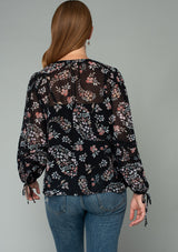 [Color: Black/Dusty Rose] A back facing image of a red headed model wearing a bohemian chiffon blouse in a black and pink floral print. With voluminous long sleeves, adjustable wrist ties, and a self covered button front. 