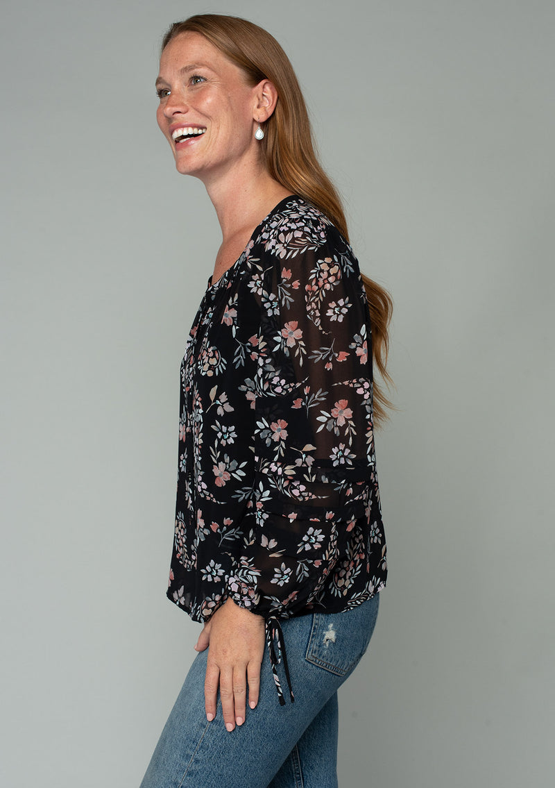 [Color: Black/Dusty Rose] A side facing image of a red headed model wearing a bohemian chiffon blouse in a black and pink floral print. With voluminous long sleeves, adjustable wrist ties, and a self covered button front. 