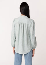 [Color: Sage] A back facing image of a brunette model wearing a classic bohemian cotton sage green button front shirt in a textured jacquard. With long sleeves and a ruffled neckline.