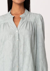 [Color: Sage] A close up front facing image of a brunette model wearing a classic bohemian cotton sage green button front shirt in a textured jacquard. With long sleeves and a ruffled neckline.