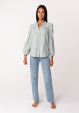 [Color: Sage] A front facing image of a brunette model wearing a classic bohemian cotton sage green button front shirt in a textured jacquard. With long sleeves and a ruffled neckline.