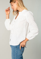 [Color: White] A side facing image of a blonde model wearing a classic bohemian cotton white button front shirt in a textured jacquard. With long sleeves and a ruffled neckline. 