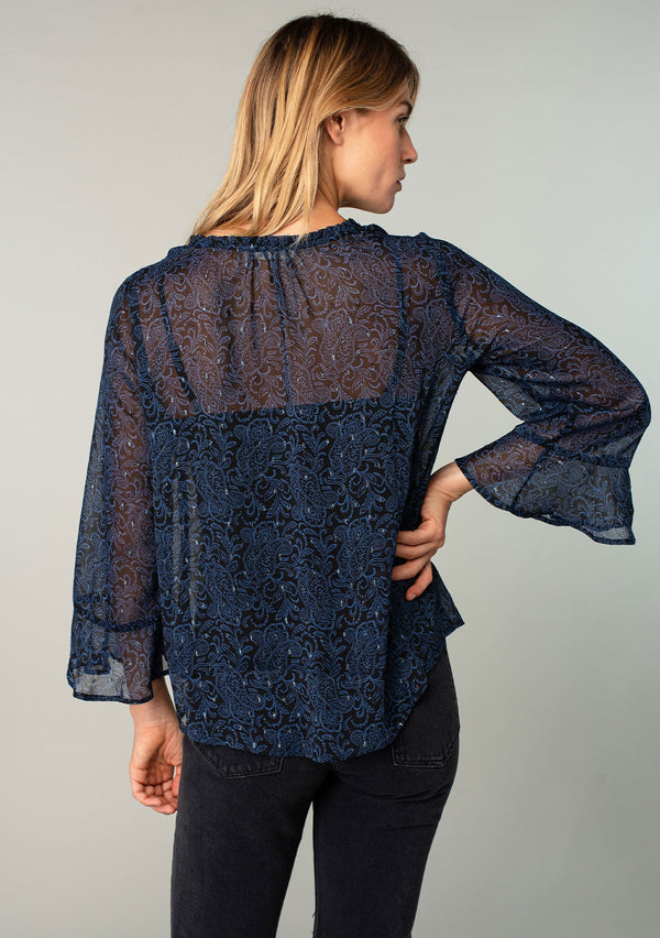 [Color: Black/Blue] A back facing image of a blonde model wearing a sheer chiffon bohemian blouse in a navy blue and black paisley print. With metallic clip dot details, three quarter length sleeves, flutter wrist cuffs, and a ruffled neckline. 