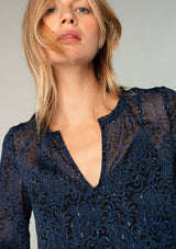 [Color: Black/Blue] A close up front facing image of a blonde model wearing a sheer chiffon bohemian blouse in a navy blue and black paisley print. With metallic clip dot details, three quarter length sleeves, flutter wrist cuffs, and a ruffled neckline. 