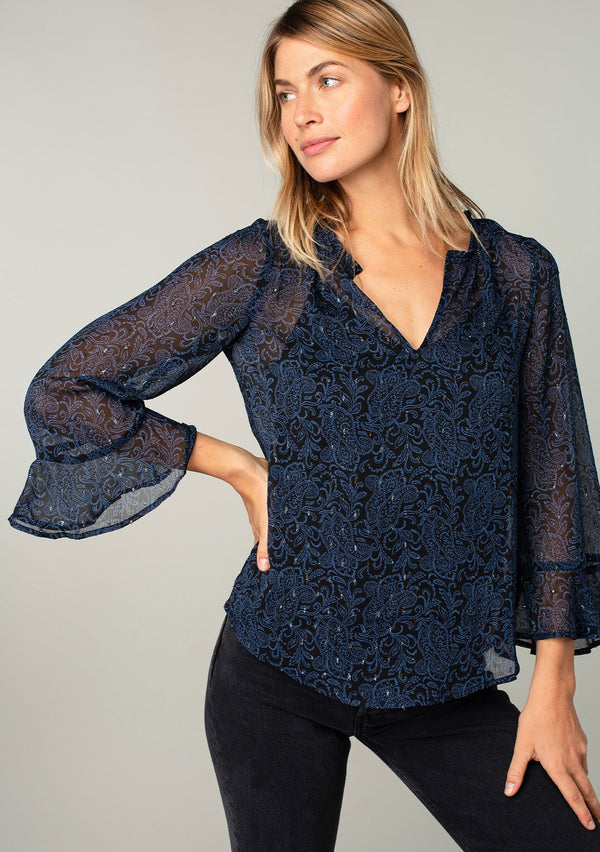 [Color: Black/Blue] A front facing image of a blonde model wearing a sheer chiffon bohemian blouse in a navy blue and black paisley print. With metallic clip dot details, three quarter length sleeves, flutter wrist cuffs, and a ruffled neckline. 