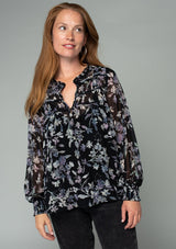 [Color: Black/Lavender] A half body front facing image of a red headed model wearing a sheer chiffon bohemian holiday blouse in a black and lavender purple floral print. With long sheer sleeves, a self covered button front, a ruffled neckline, and a vintage inspired front yoke detail. 