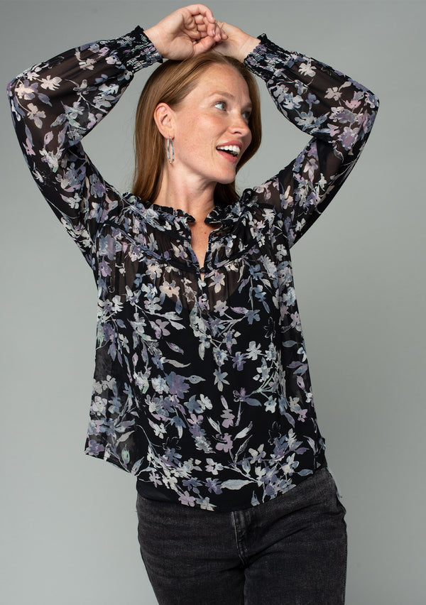 [Color: Black/Lavender] A front facing image of a red headed model wearing a sheer chiffon bohemian holiday blouse in a black and lavender purple floral print. With long sheer sleeves, a self covered button front, a ruffled neckline, and a vintage inspired front yoke detail. 