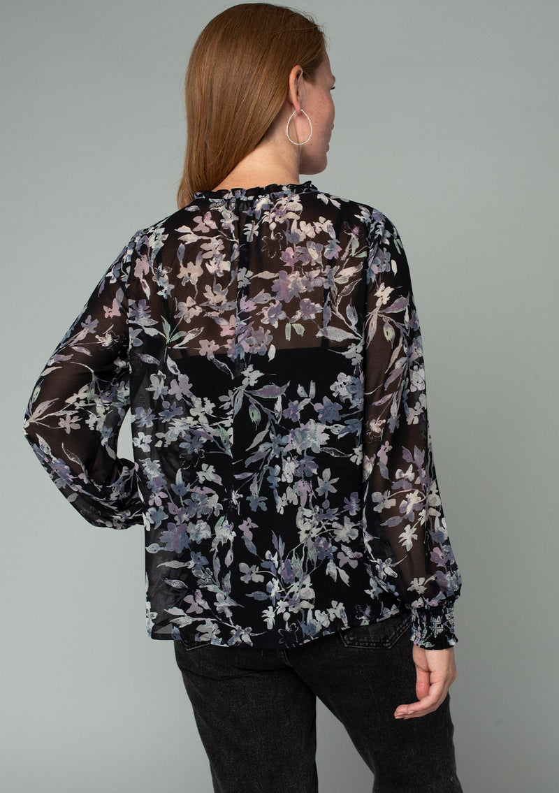 [Color: Black/Lavender] A back facing image of a red headed model wearing a sheer chiffon bohemian holiday blouse in a black and lavender purple floral print. With long sheer sleeves, a self covered button front, a ruffled neckline, and a vintage inspired front yoke detail. 