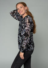 [Color: Black/Lavender] A side facing image of a red headed model wearing a sheer chiffon bohemian holiday blouse in a black and lavender purple floral print. With long sheer sleeves, a self covered button front, a ruffled neckline, and a vintage inspired front yoke detail. 