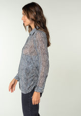 [Color: Grey/Natural] A side facing image of a brunette model wearing a sheer chiffon button front shirt in a grey and natural paisley print. With a collared neckline, long sleeves with a button wrist cuff, and an adjustable side gathered waist detail with ties. 