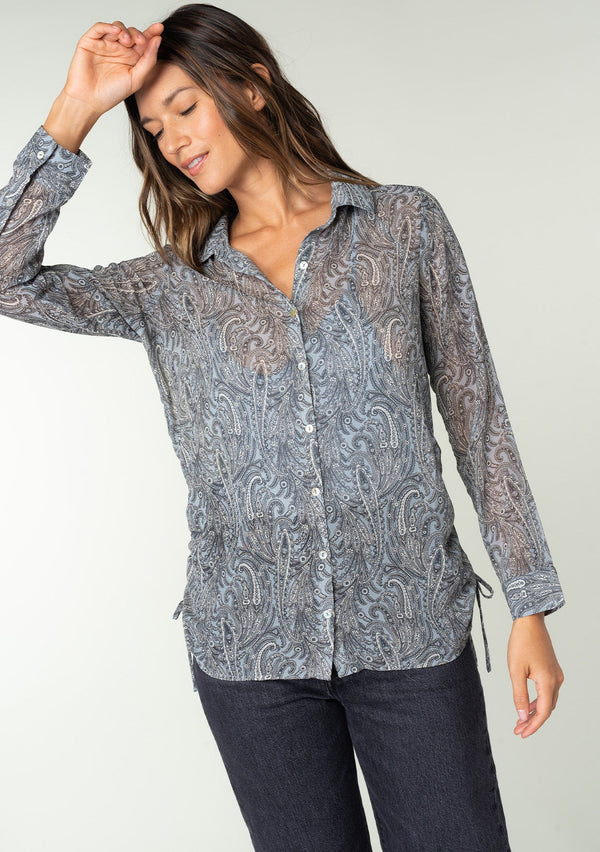 [Color: Grey/Natural] A front facing image of a brunette model wearing a sheer chiffon button front shirt in a grey and natural paisley print. With a collared neckline, long sleeves with a button wrist cuff, and an adjustable side gathered waist detail with ties. 