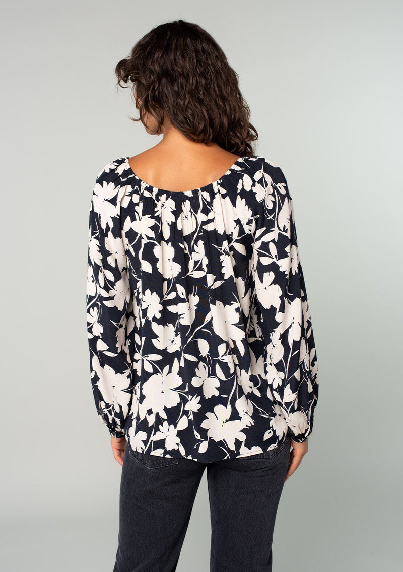 [Color: Black/Natural] A back facing image of a brunette model wearing a black and off white floral print blouse. A bohemian resort top with long sleeves and a wide elastic neckline. 